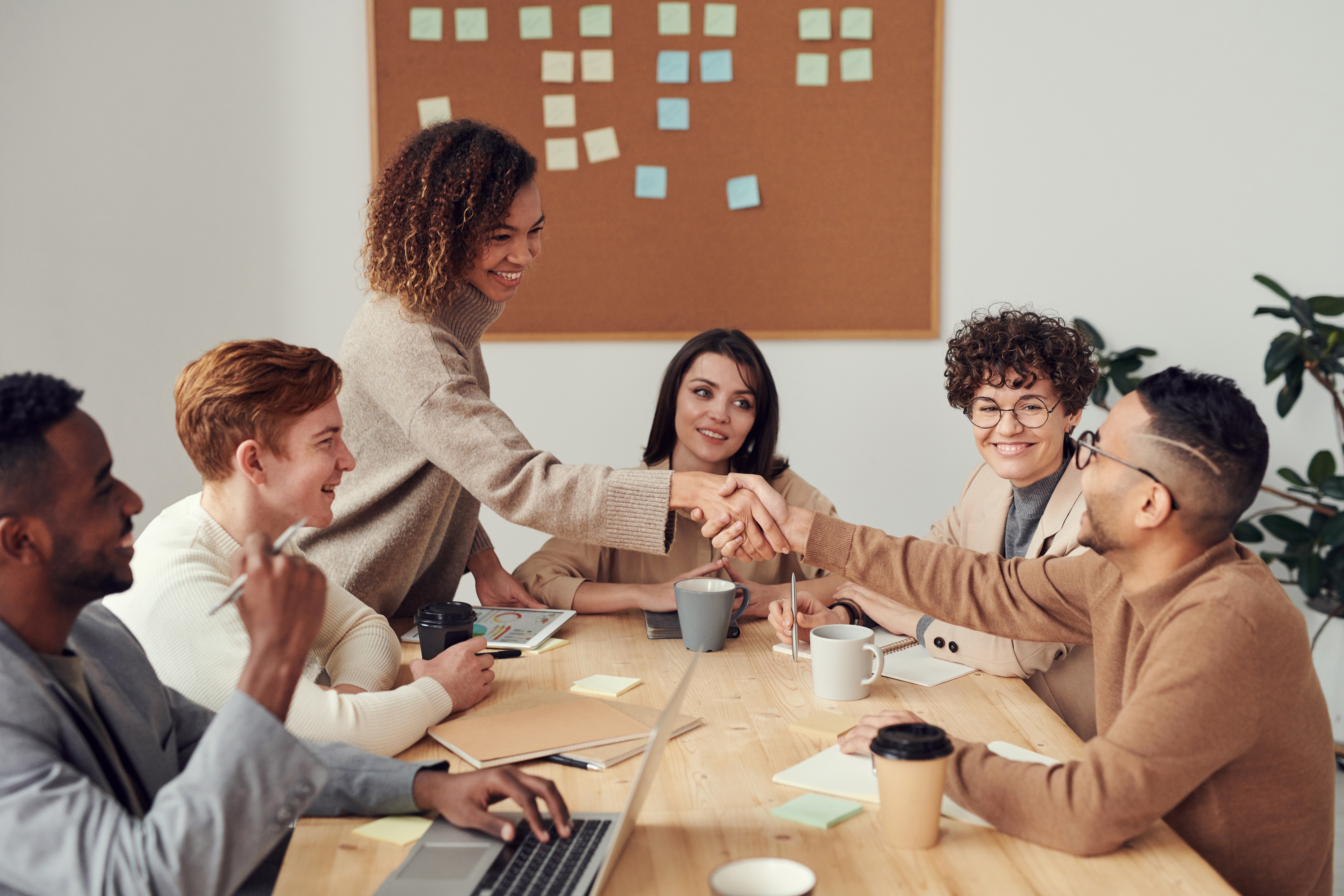 Two people shaking hands at a table with other smiling people. Backdrop of a corkboard with sticky notes on it.