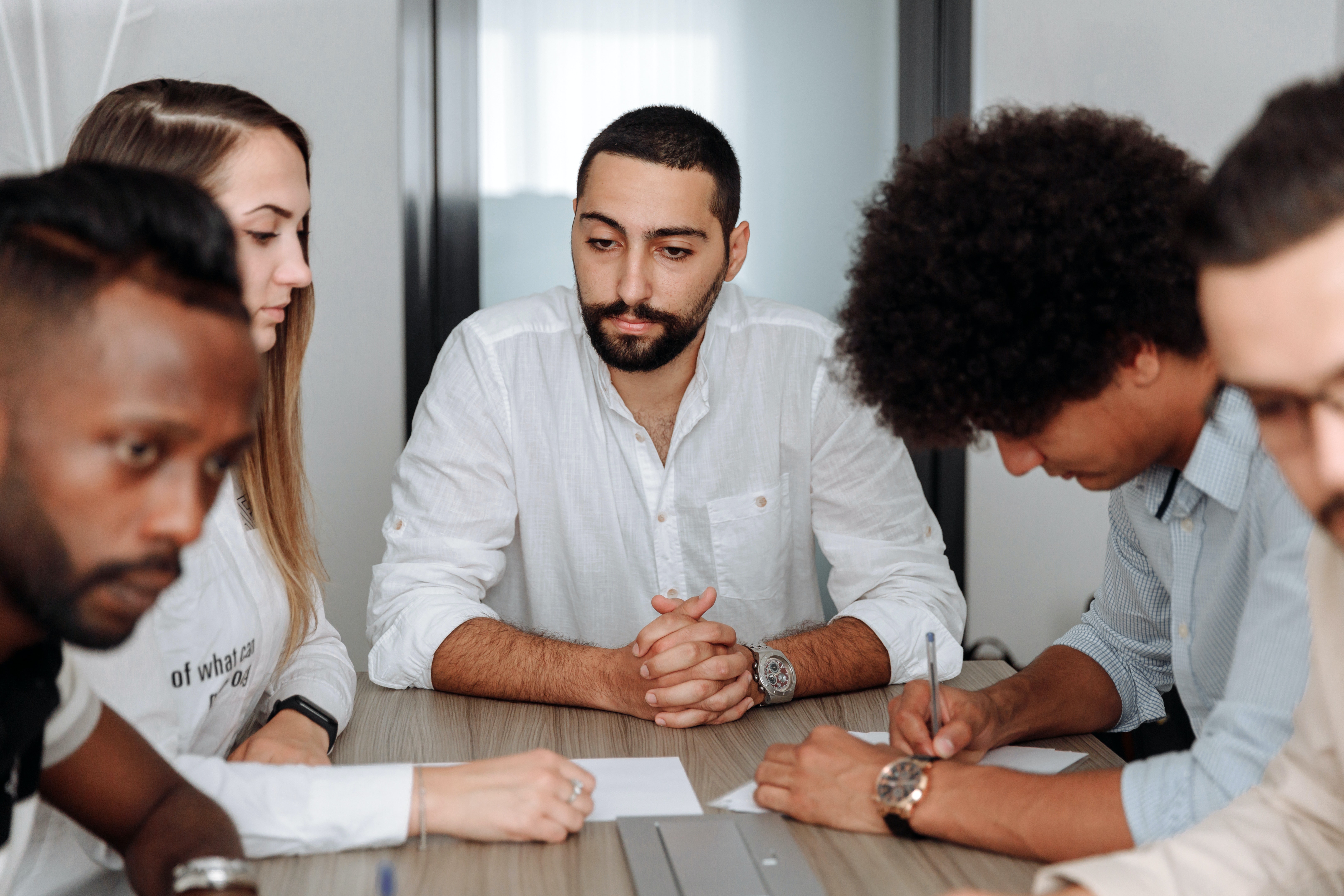 Group of people at a table taking notes. One man in the center appears to be thinking.